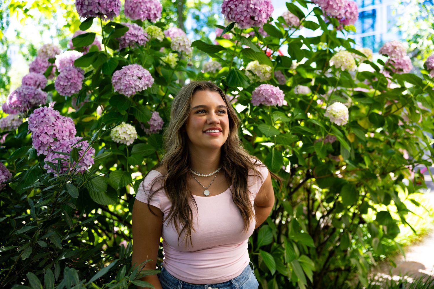 A pink tank top wearer sits in front of vibrant purple and pink hydrangea bushes on a sunny summer day.
