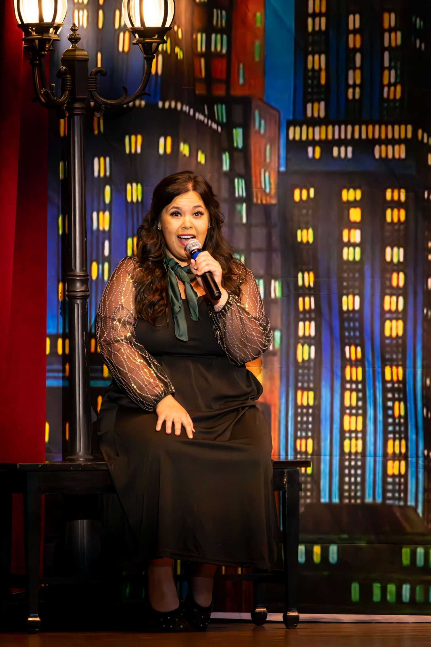 A performer in a black dress sits on stage during a night-time talk show segment with a city backdrop.