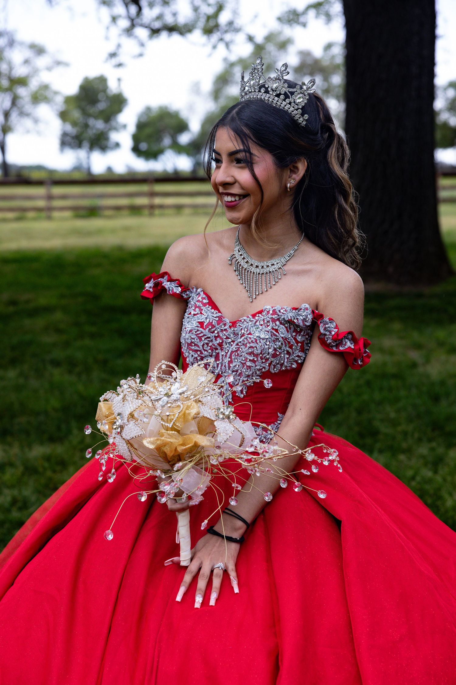 A red quinceañera dress with crystal embellishments and off-shoulder design is showcased in an outdoor garden setting.
