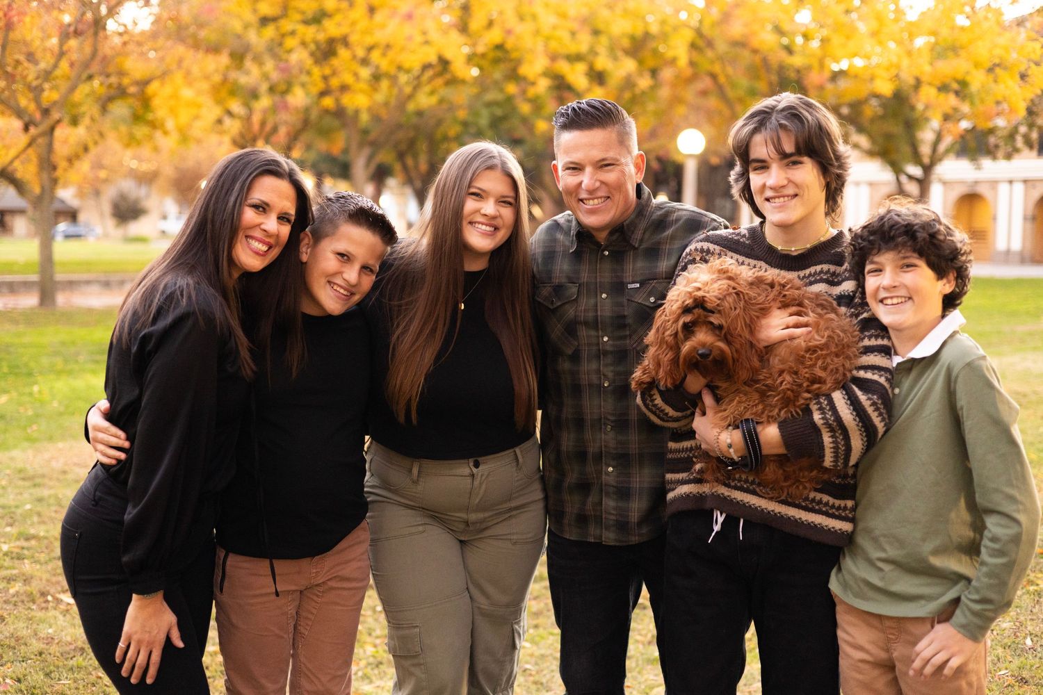 A group stands together outdoors on a sunny autumn day with golden leaves in the background.