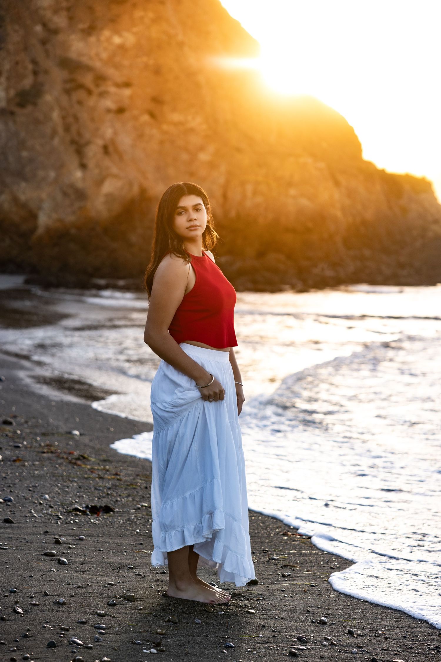A person in a red top and flowing skirt stands on a beach at sunset with cliffs in the background in Pacifica.