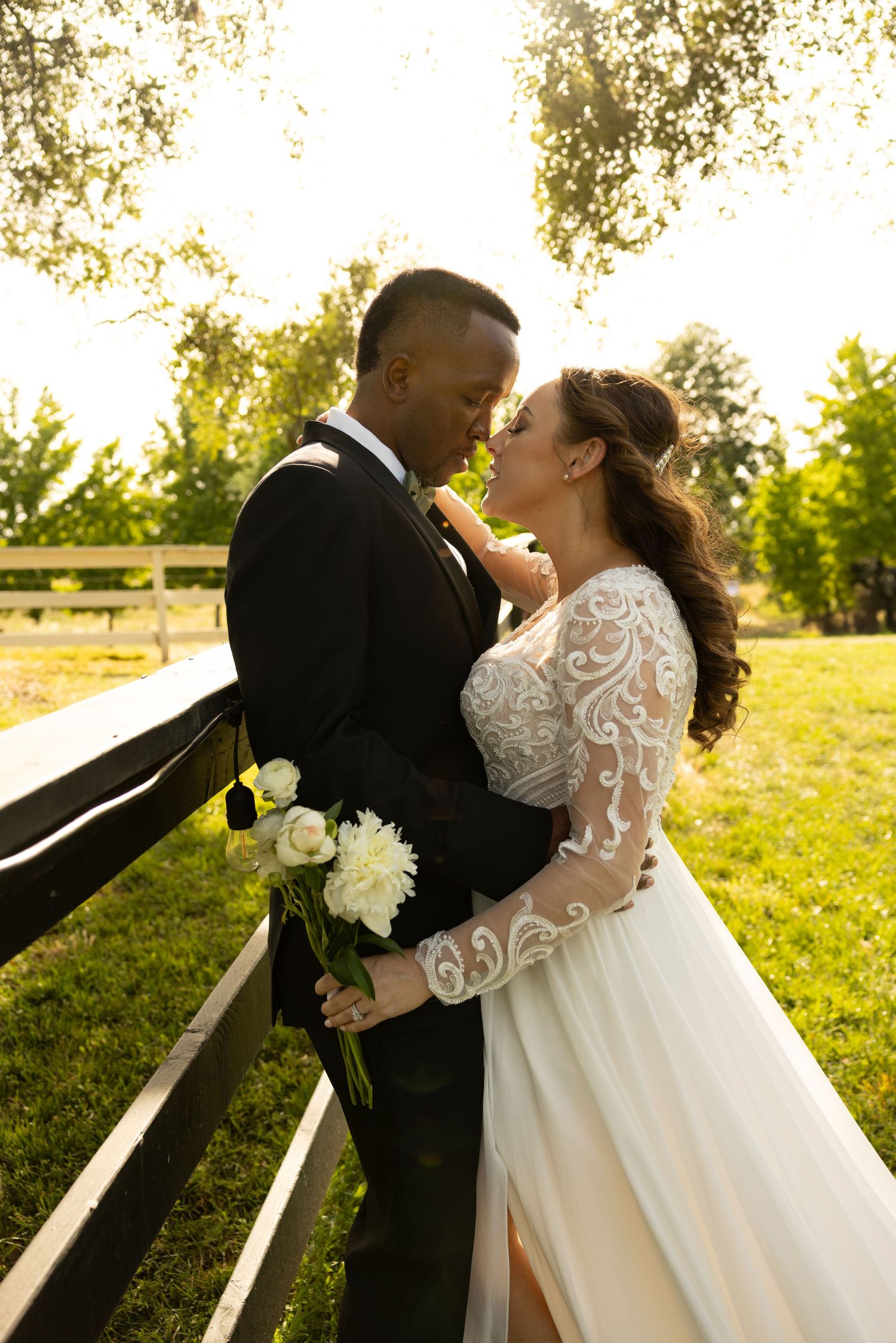 Couple in wedding attire share an intimate moment on wooden steps in a sunlit outdoor setting, holding white flowers.