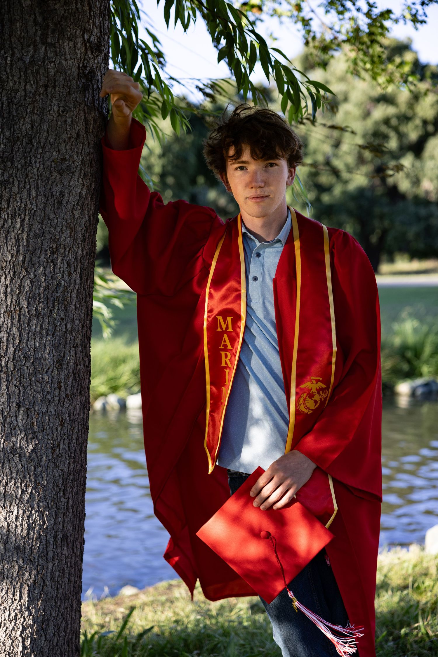 Student in red graduation gown poses for portraits by a lake on a sunny day.