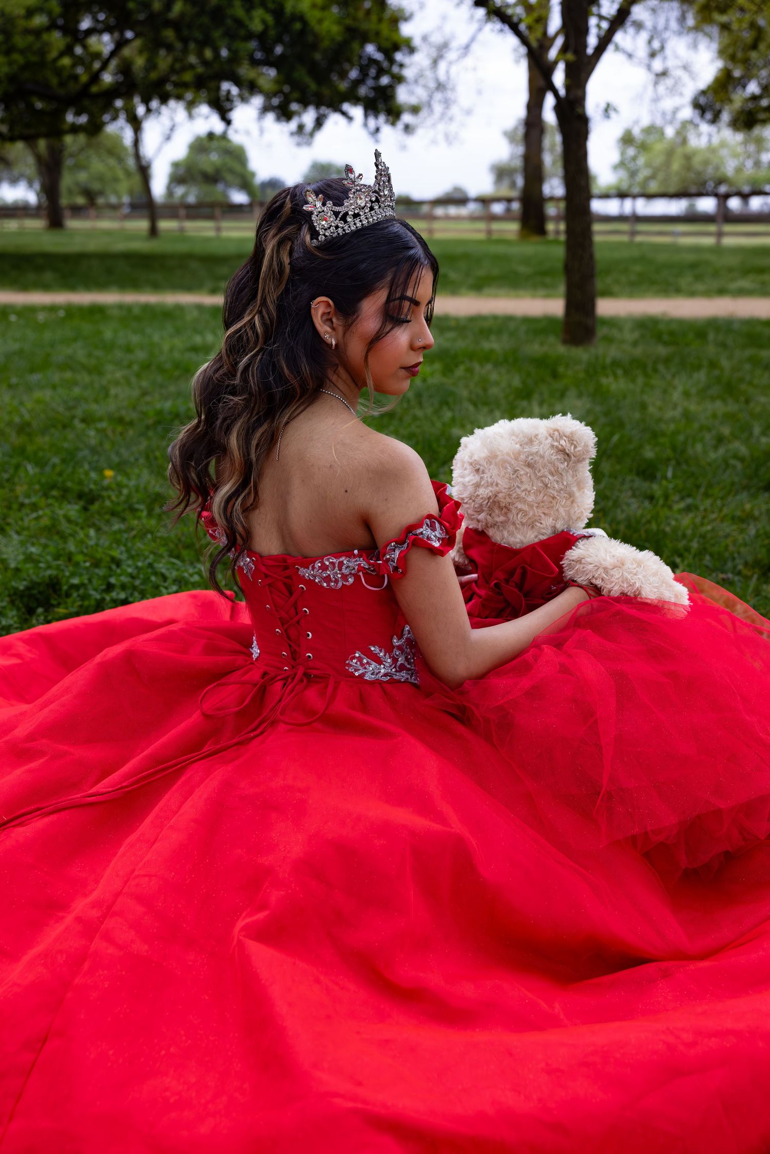 A person in a flowing red quinceañera dress holds a teddy bear while sitting in a park setting.