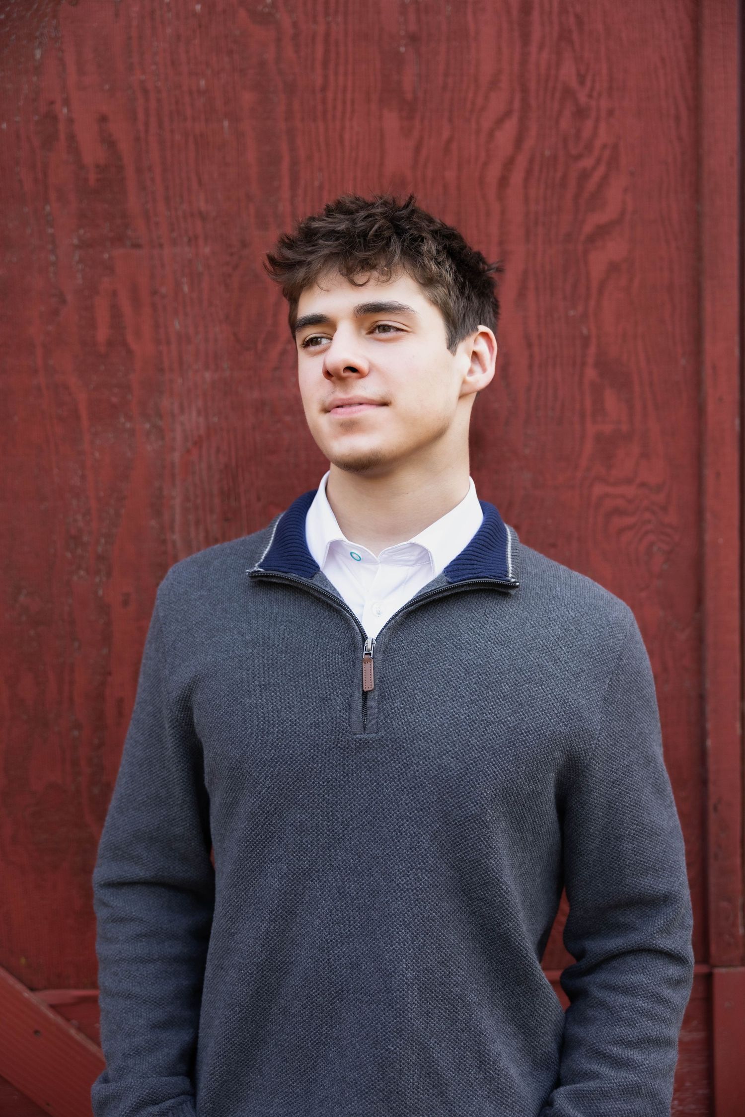 Senior Portrait against red barn wall wearing grey quarter-zip sweater and looking into the future in Chico, California