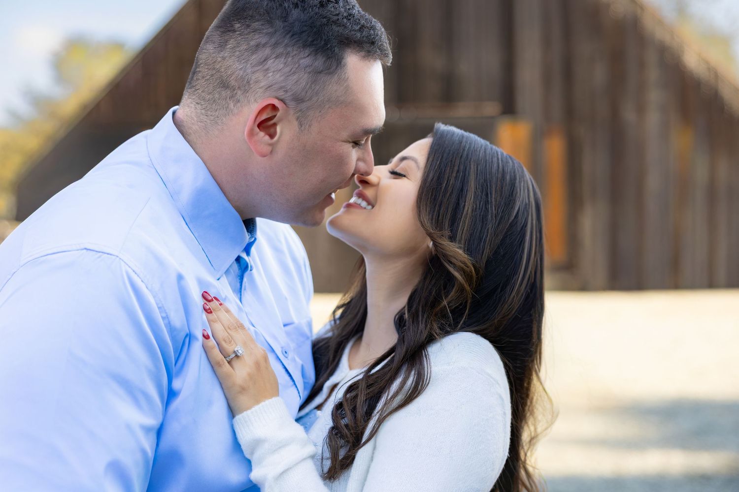 A couple in coordinated blue and white outfits share a romantic moment outdoors in a rustic setting.