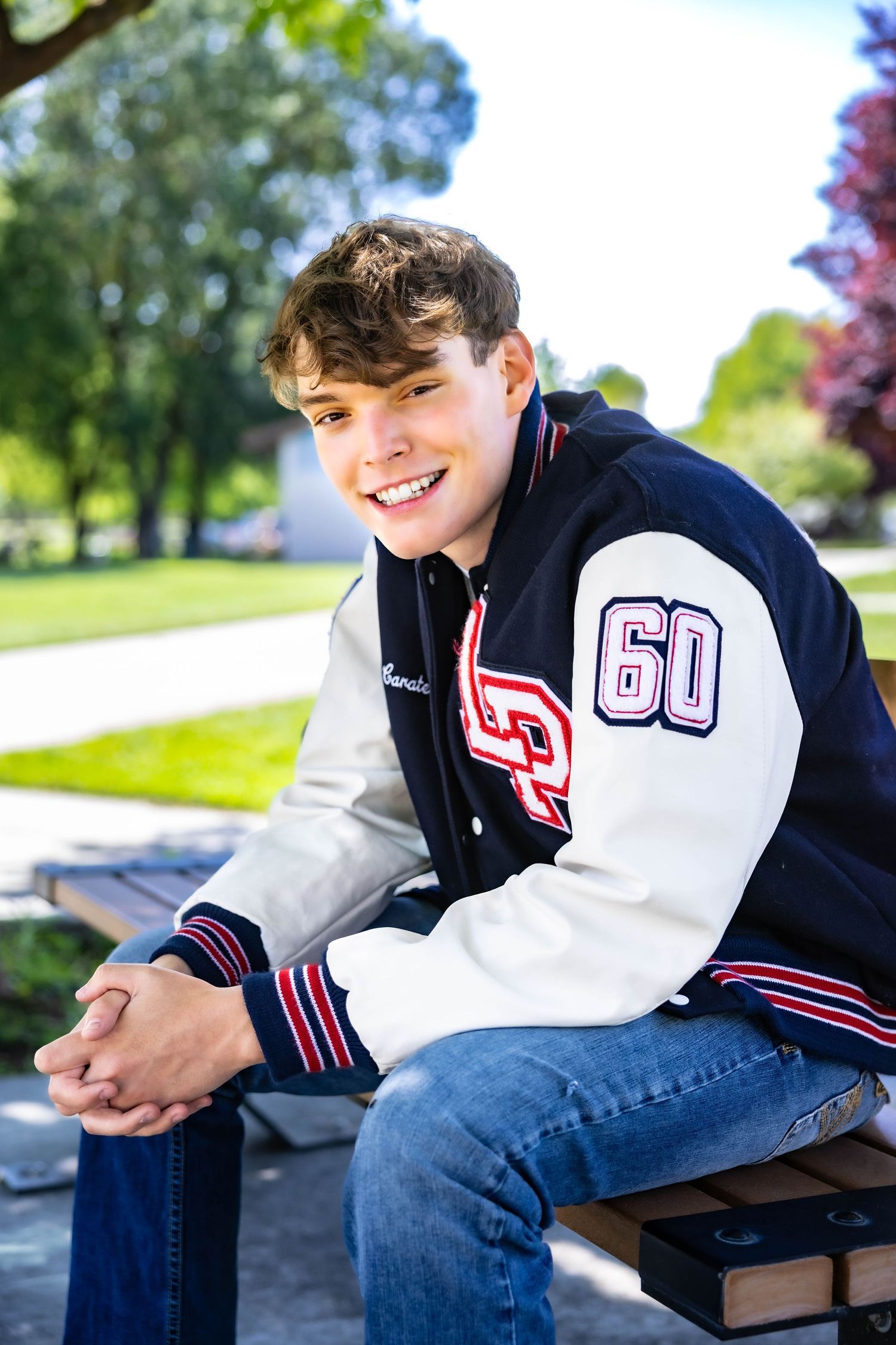 Senior football player in navy and white varsity jacket sits on bench in sunny park setting with trees in background.