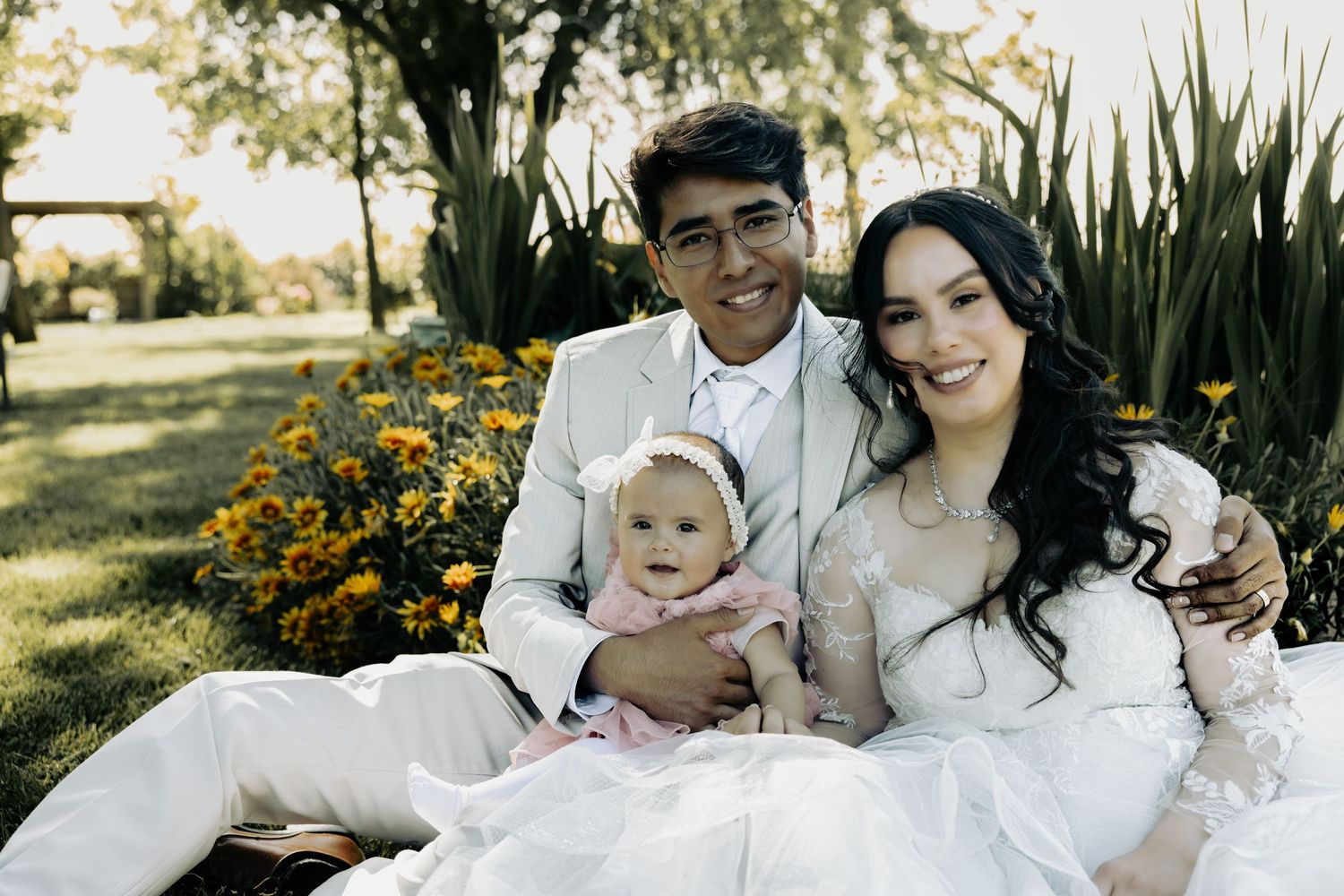 A happy couple in formal wedding attire sitting outdoors with their baby in a garden setting with yellow flowers.