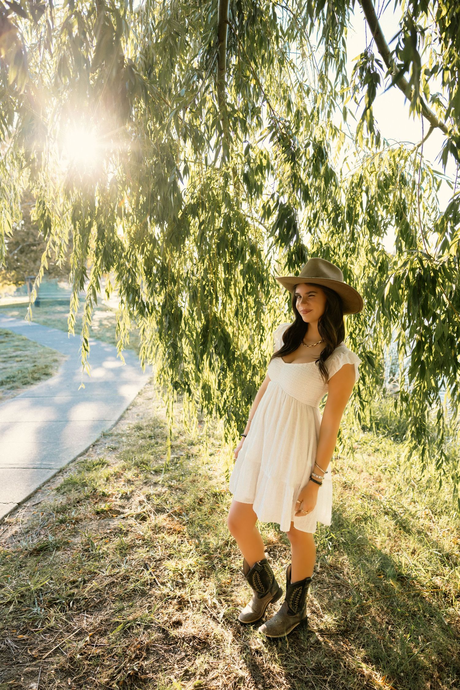 A figure in a white dress and sun hat stands beneath drooping willow branches in golden afternoon sunlight.