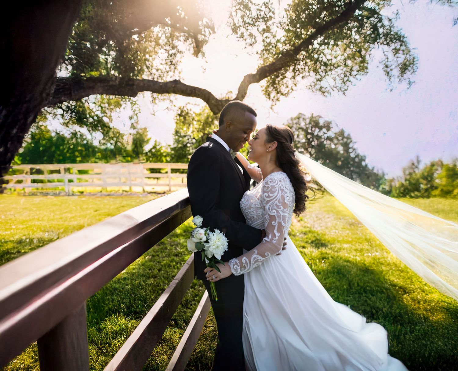 A couple in wedding attire embraces on a rustic wooden bridge during a golden sunset in a countryside setting.