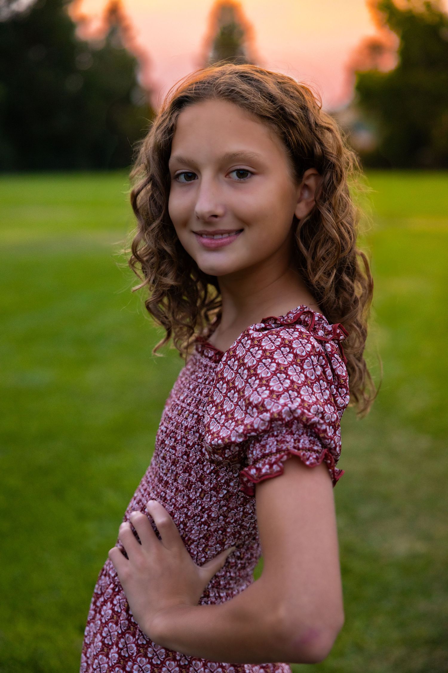 A person in a red floral dress poses in a green field at sunset.