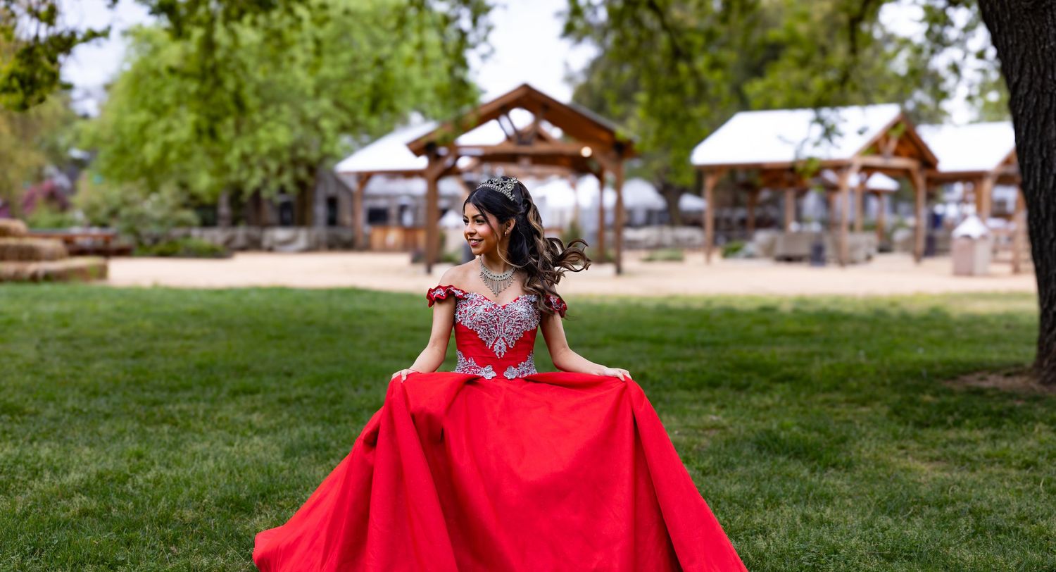 A person in a flowing red formal gown poses on a green lawn with rustic wooden pavilions in the background.