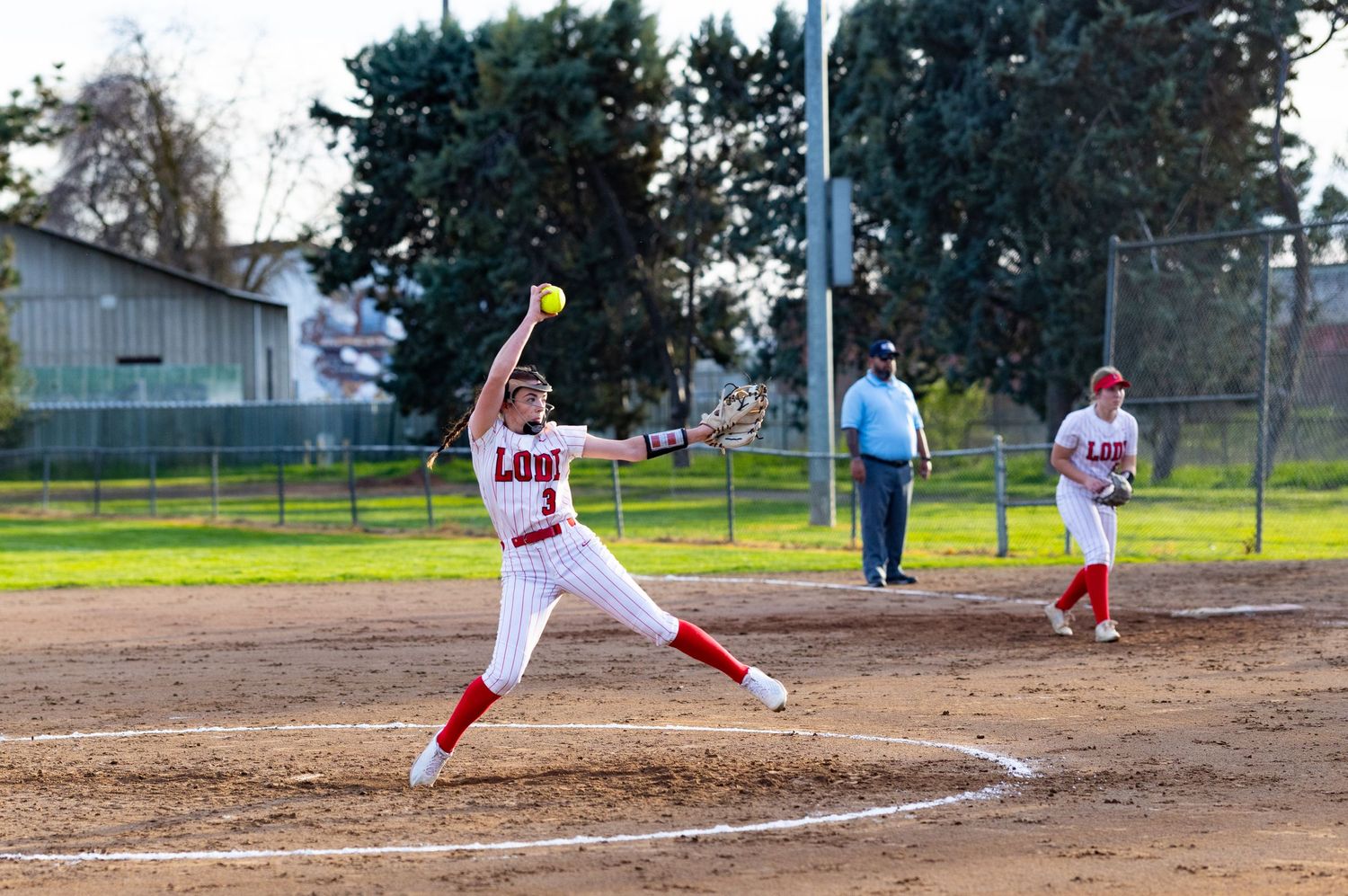 Softball pitcher Lodi High, Sports Photographer, in white and red uniform mid-throw during game.