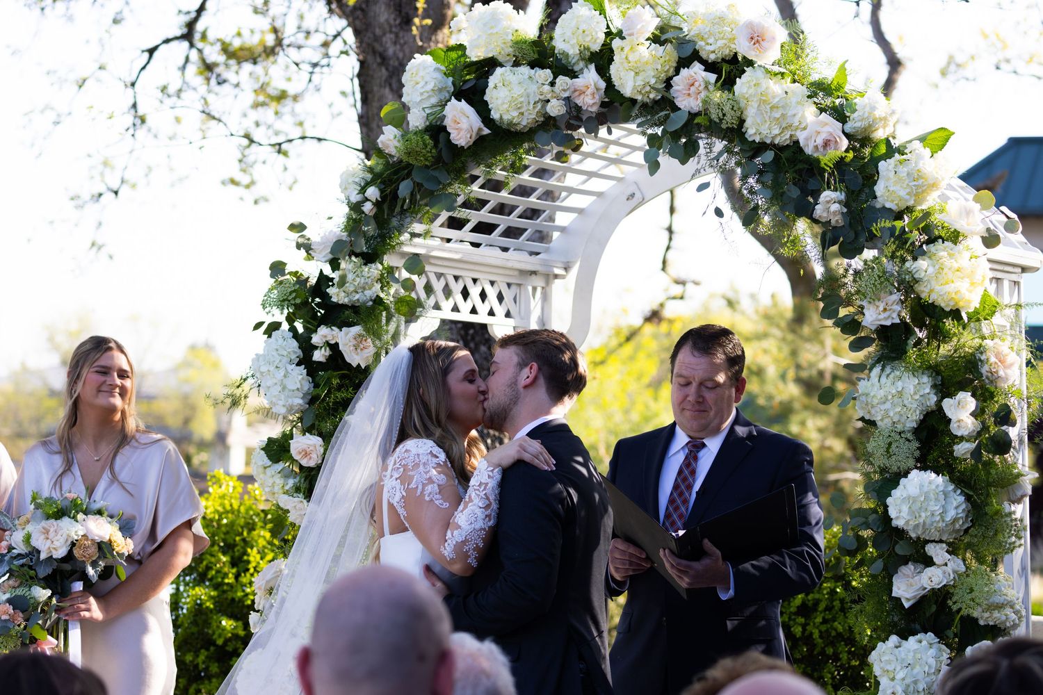 A wedding ceremony takes place under a beautiful white floral arch with lush greenery in an outdoor garden setting.