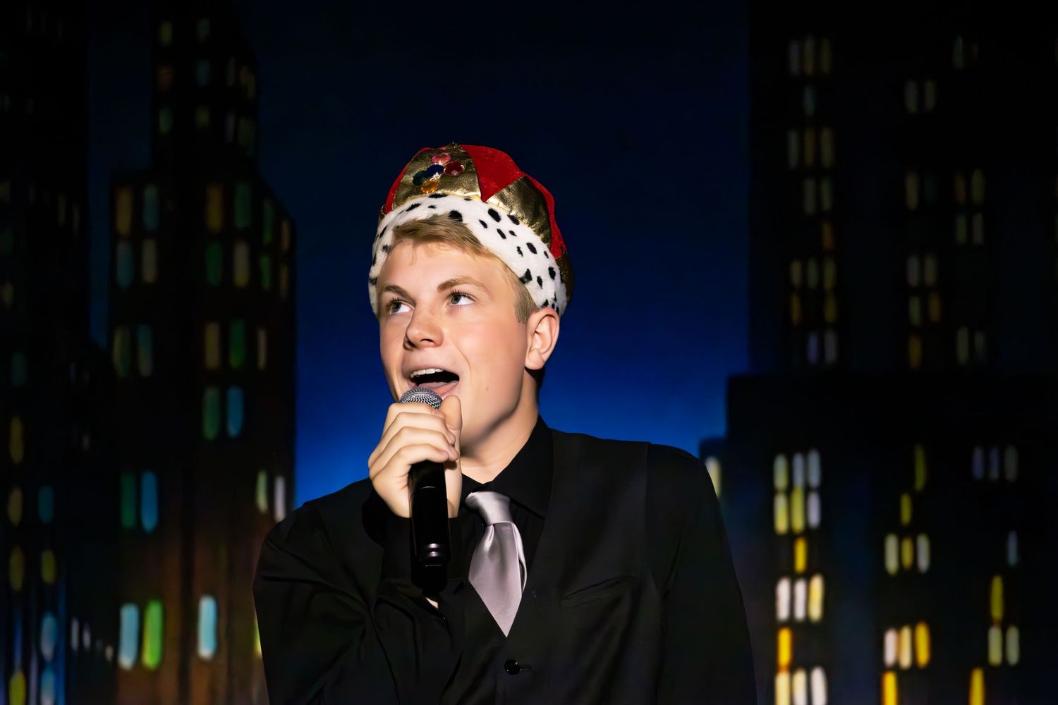 Singer performing on stage against backdrop of illuminated city skyline wearing black suit and polka dot headband.