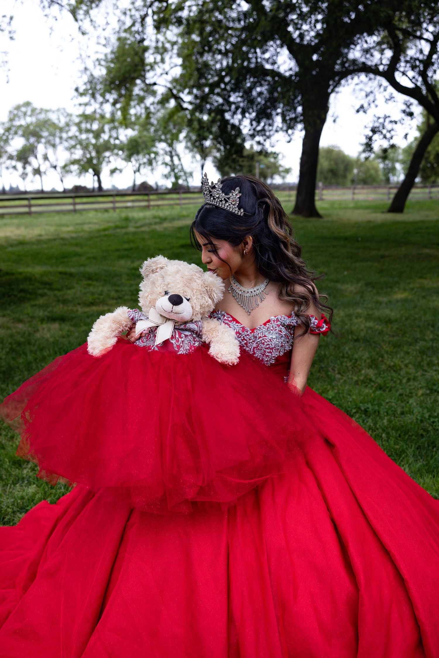 A person in a red ballgown poses with a teddy bear in a grassy outdoor setting.