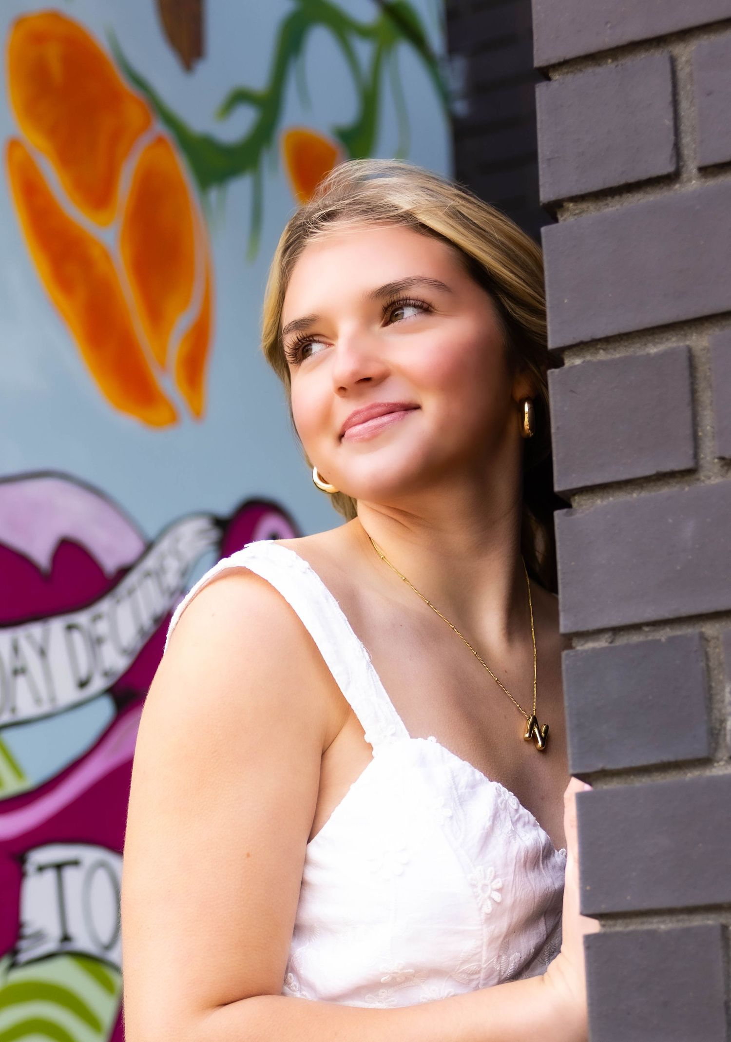 A young woman in a white sleeveless top smiles while leaning against a wall with colorful graffiti art in the background.