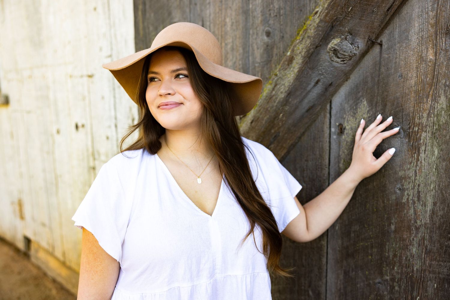 Woman in white top and brown hat poses against rustic wooden wall in natural lighting.