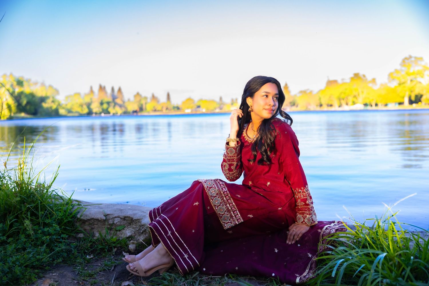 A person in a red sweater sits by a serene lakeside during golden hour with autumn trees in the background.