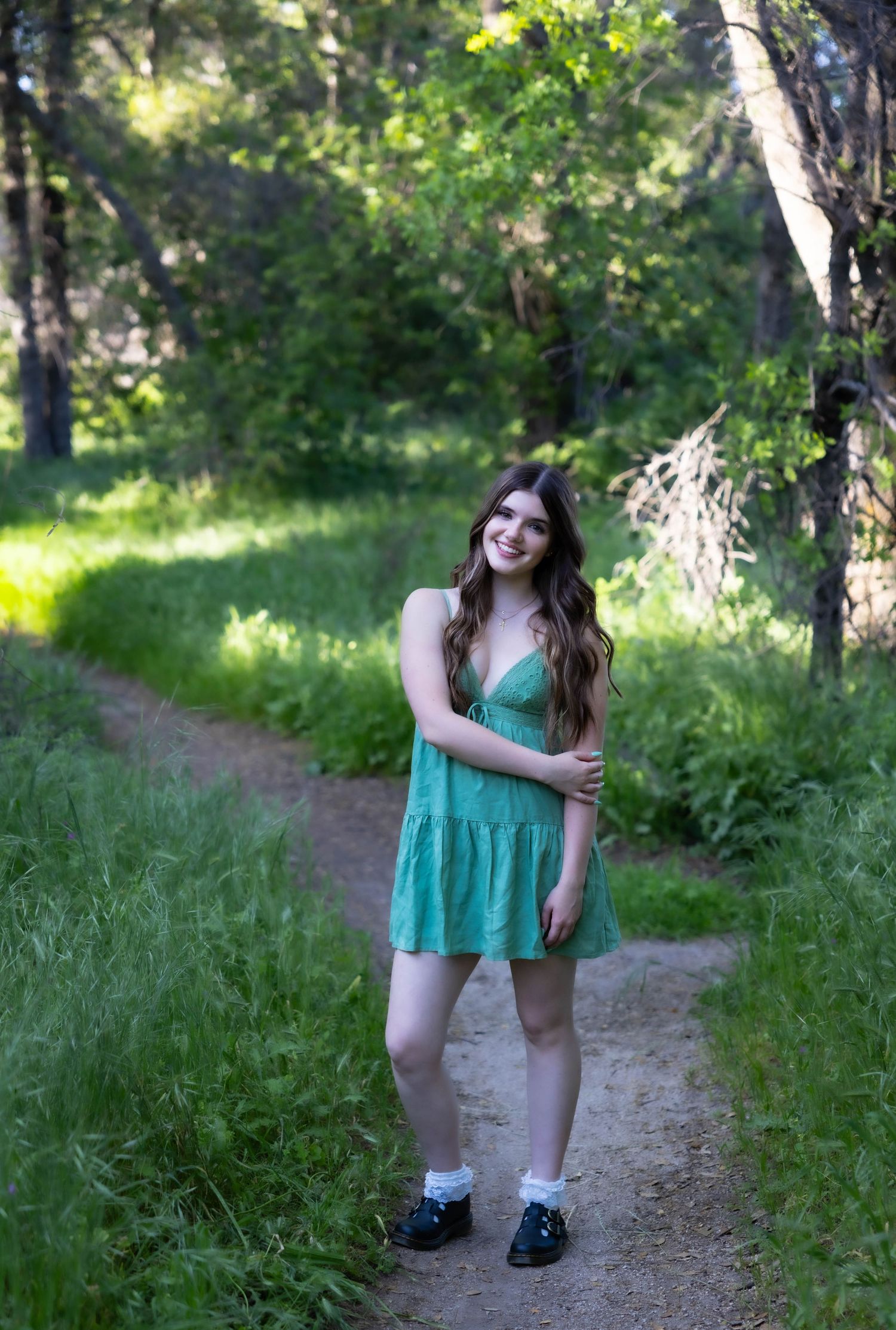 A person in a turquoise summer dress stands on a dirt path surrounded by lush green forest on a sunny day.