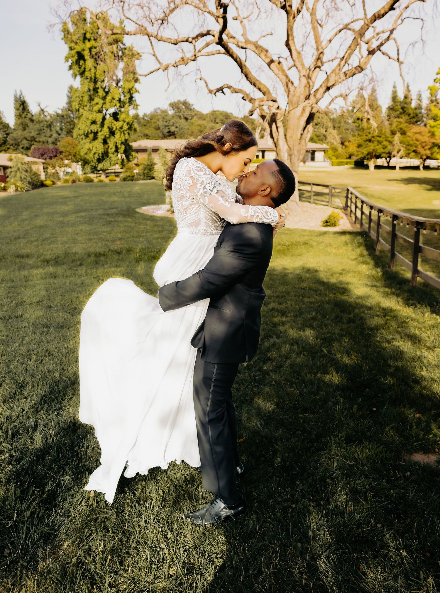 A couple in wedding attire embrace on a lush green lawn with a leafless tree and evergreens in the background.