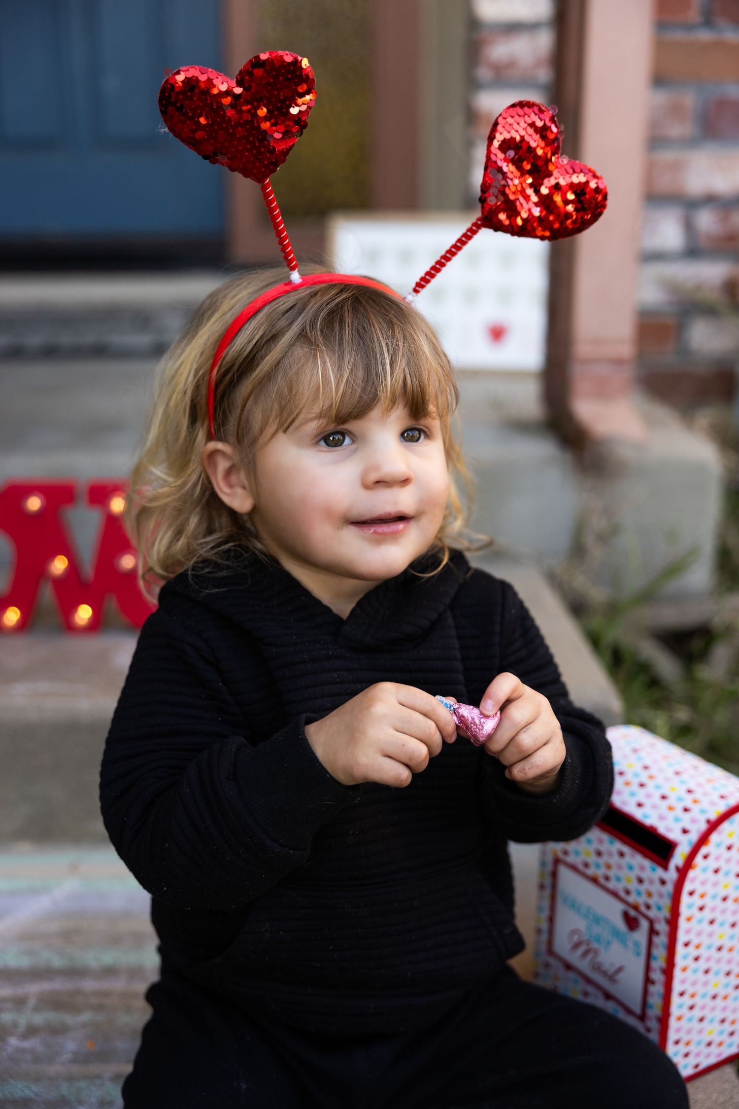 Small child wearing heart antennae and black sweater sits near Valentine's Day decorations.