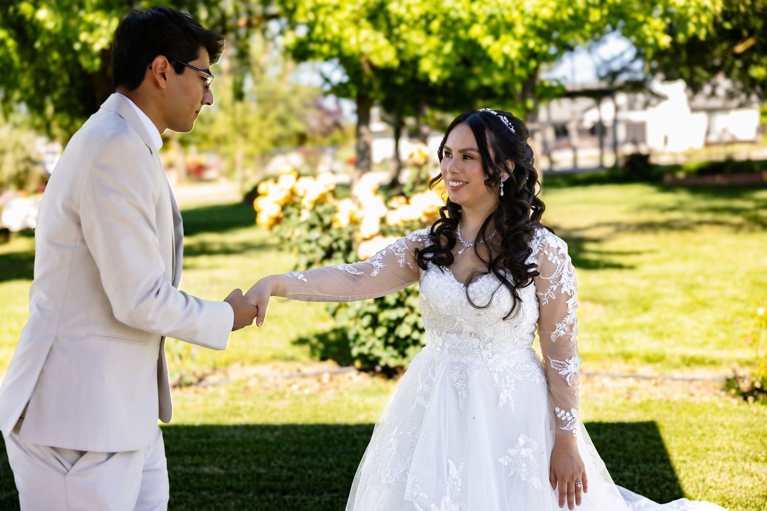 A couple in white attire walking and embracing in a sunny garden setting with green grass and trees in the background.