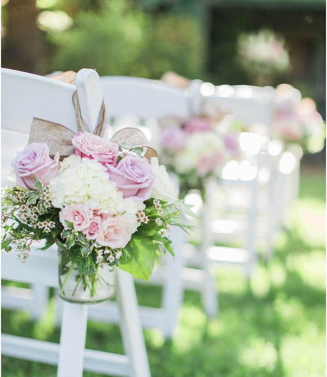 Pink and white floral arrangement with roses and hydrangeas decorates a white ceremony chair in an outdoor garden setting.
