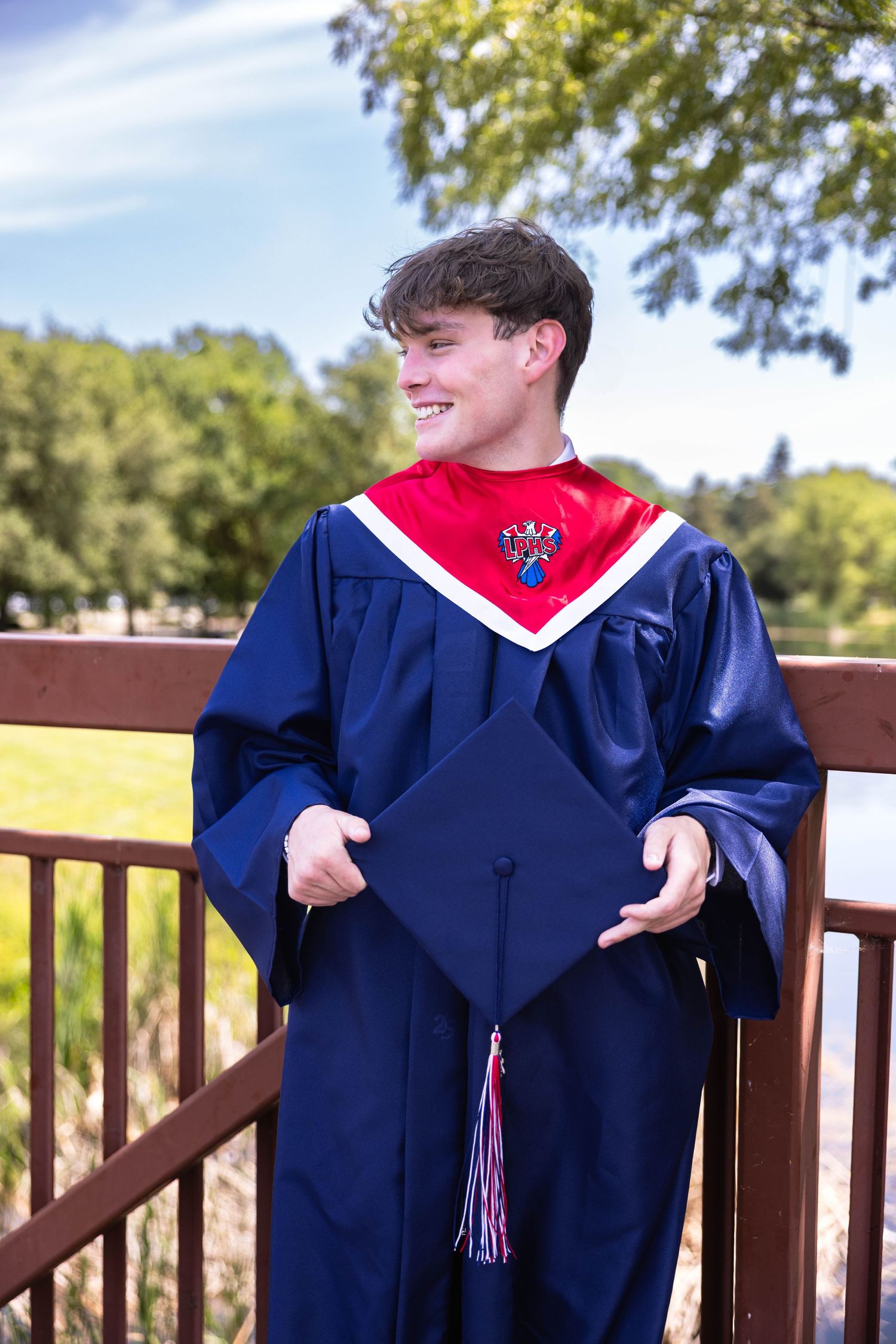 Senior poses in cap and gown on a bridge in a park for senior photos near Sacramento and Lodi.  Lake and water fountain close by, big smile
