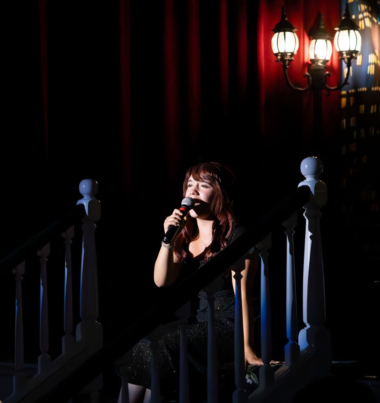 Dramatic stage performance featuring a vocalist in dark lighting with spotlights and illuminated columns visible behind.