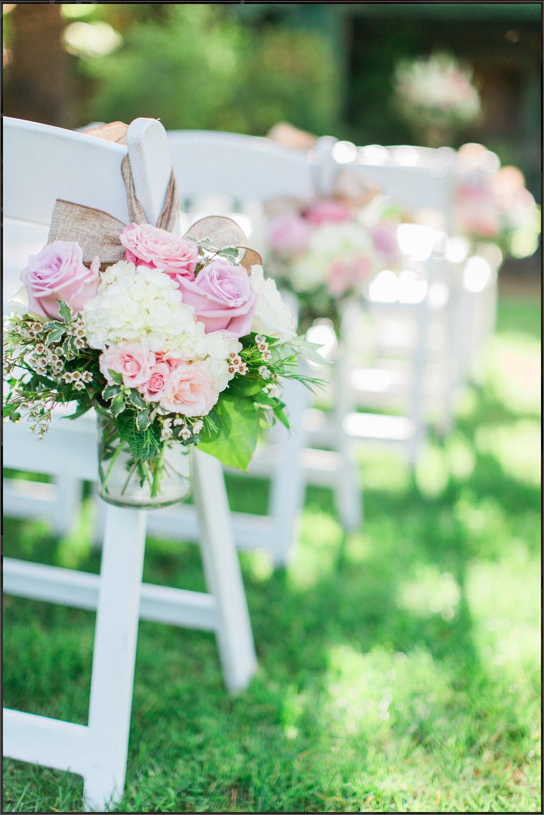 Delicate pink and white roses with greenery decorate white chairs along a grassy outdoor wedding aisle.