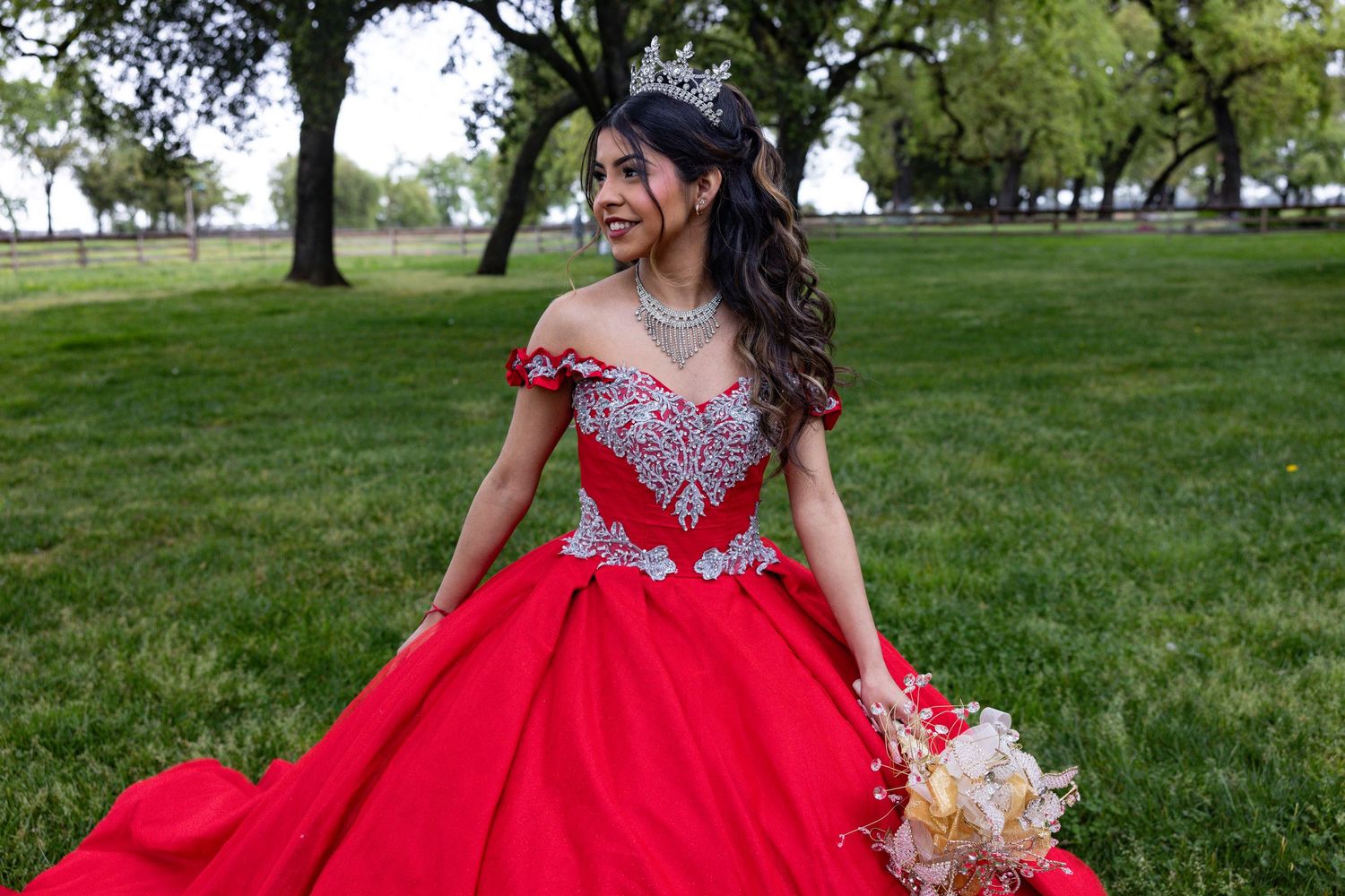 A flowing red ballgown with beaded bodice and off-shoulder details in a scenic outdoor garden setting in Sacramento.