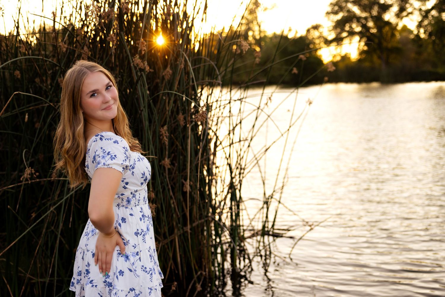 A woman in a floral dress poses by a lake at sunset among tall reeds.