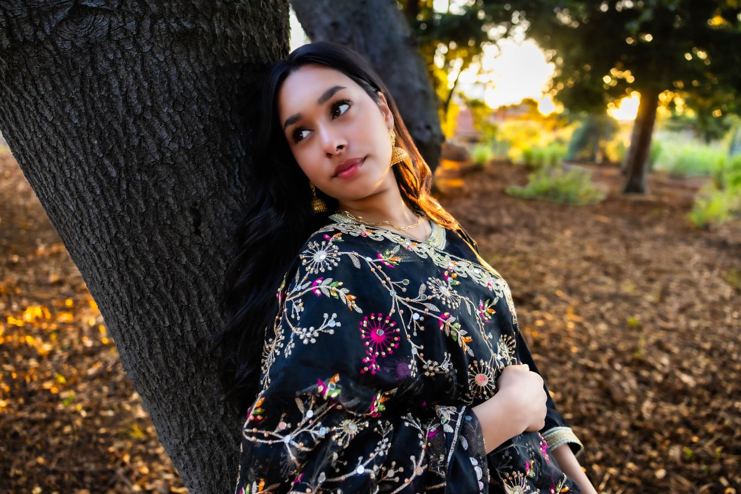A floral dress-clad figure poses gracefully against autumn trees during golden hour lighting.