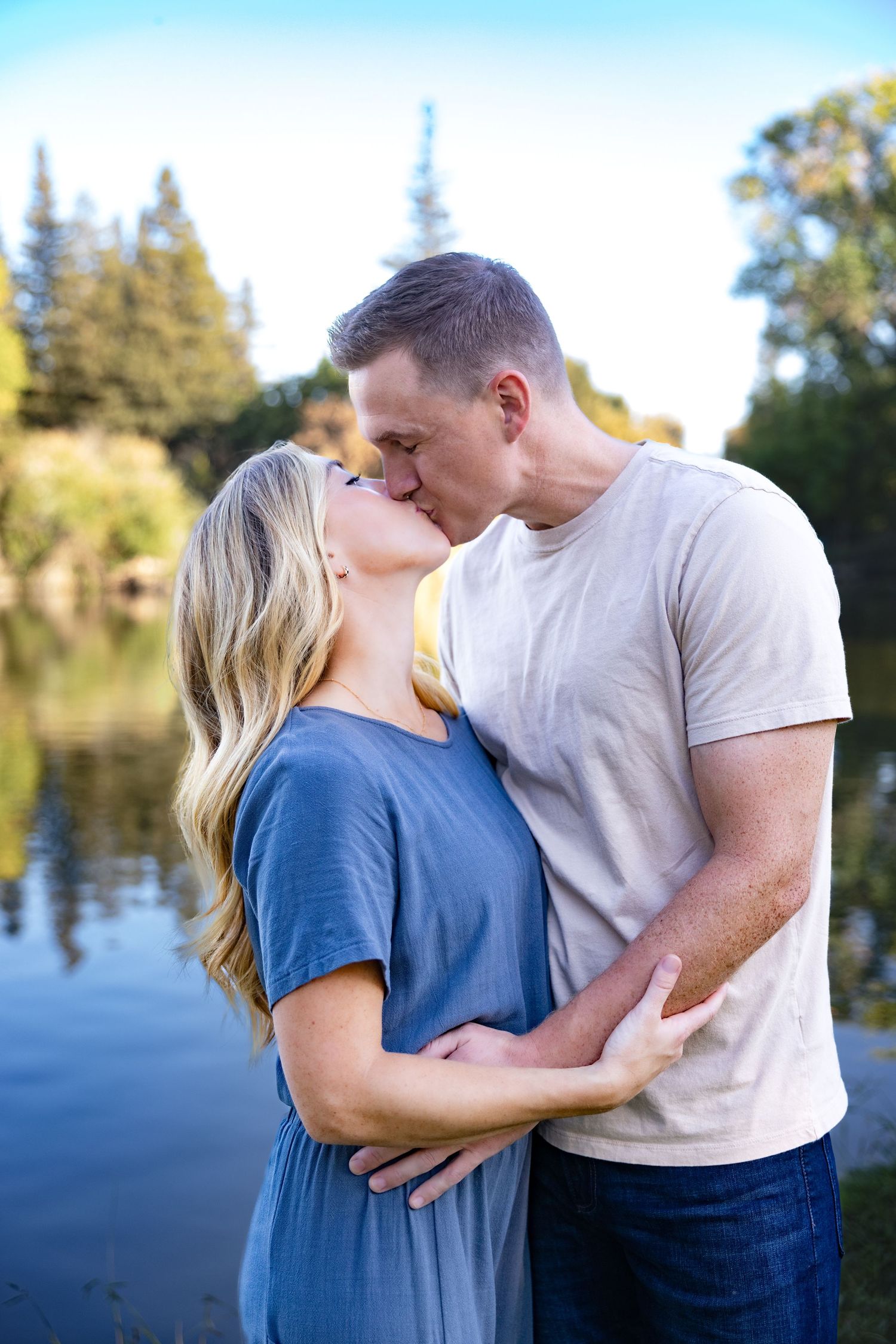 A couple in blue and white clothing shares romantic moments by a lake with trees in the background.