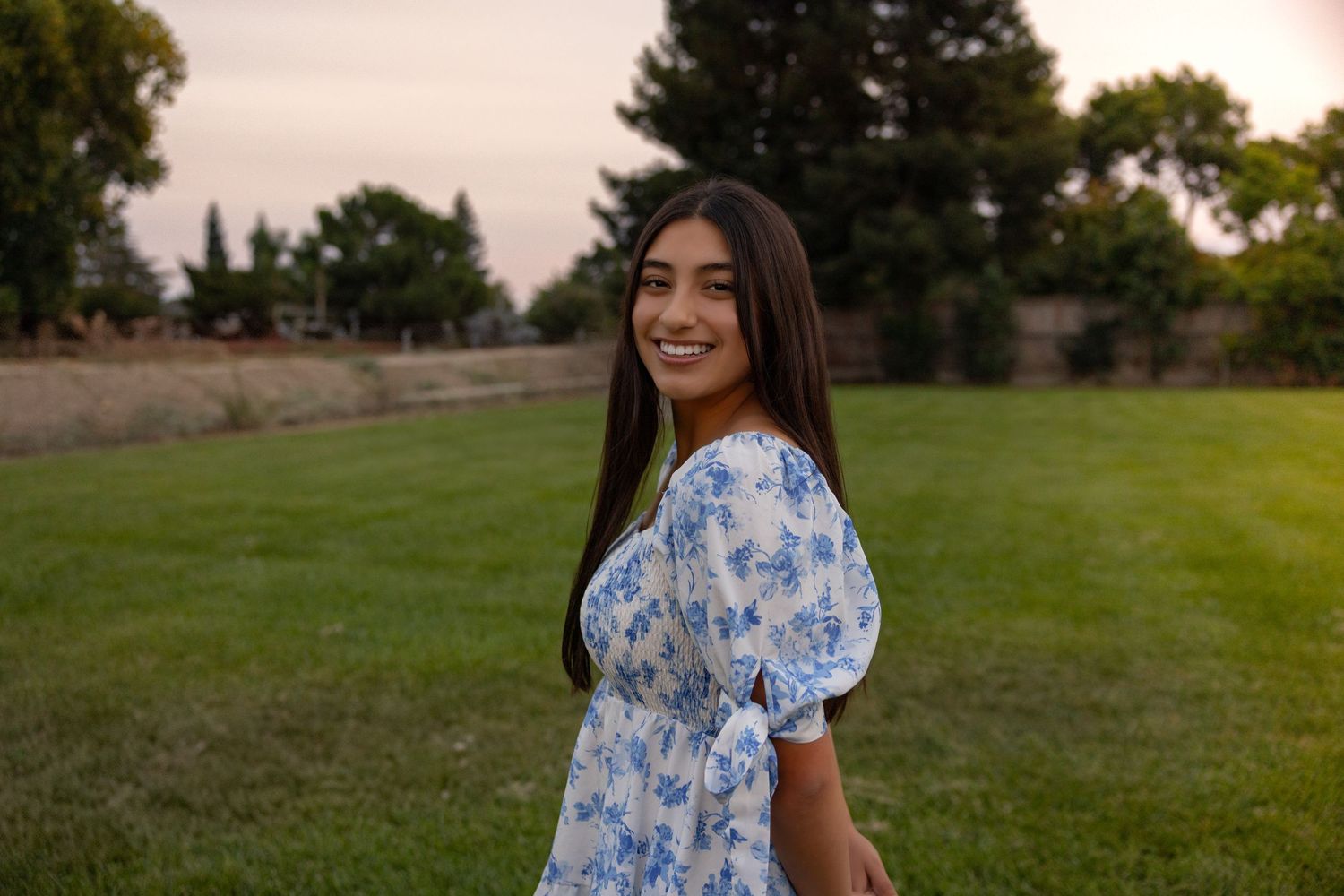 A person in a blue floral dress stands in a grassy field during golden hour.