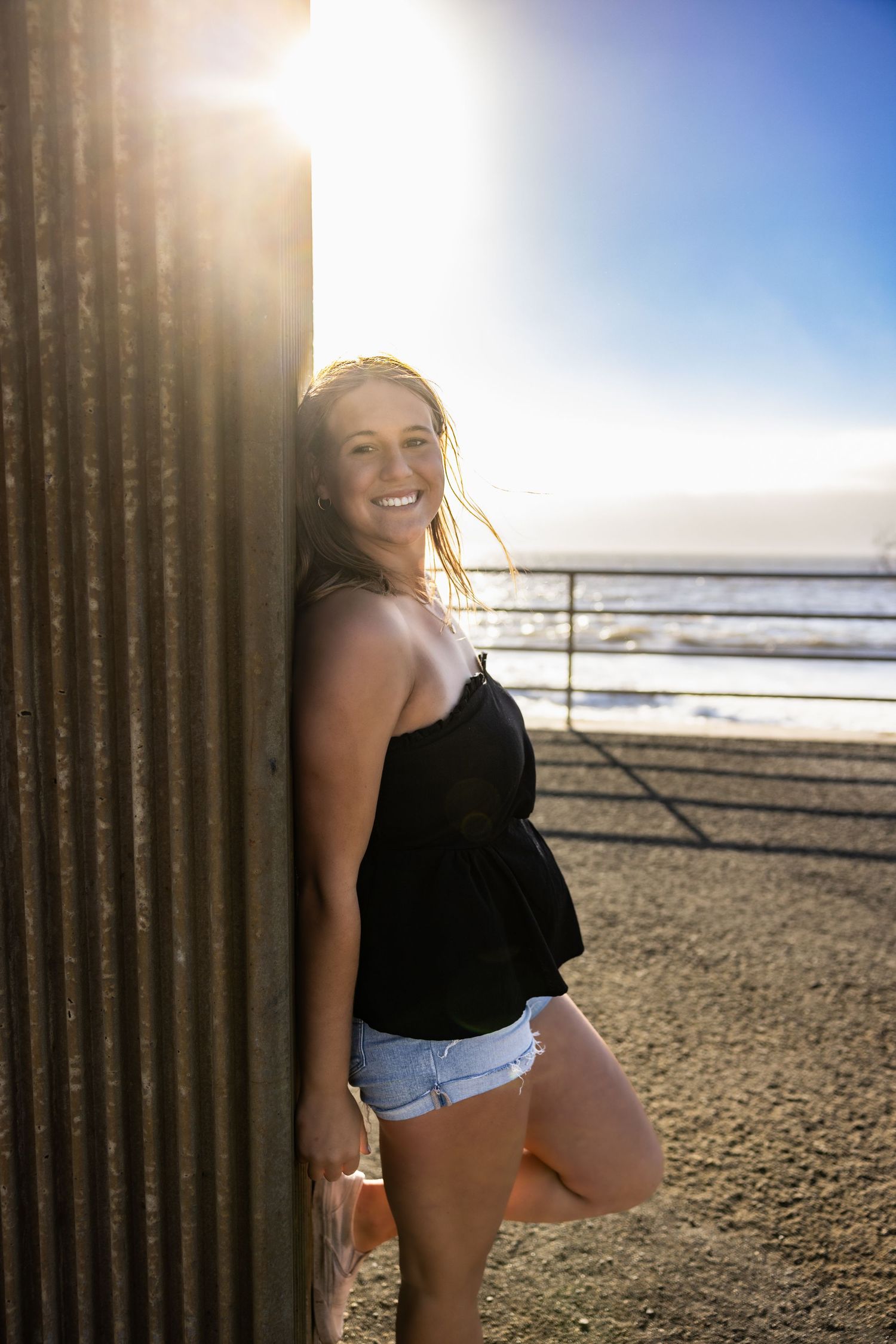 A person in casual summer attire poses against a seaside backdrop with bright sunlight and ocean waves.