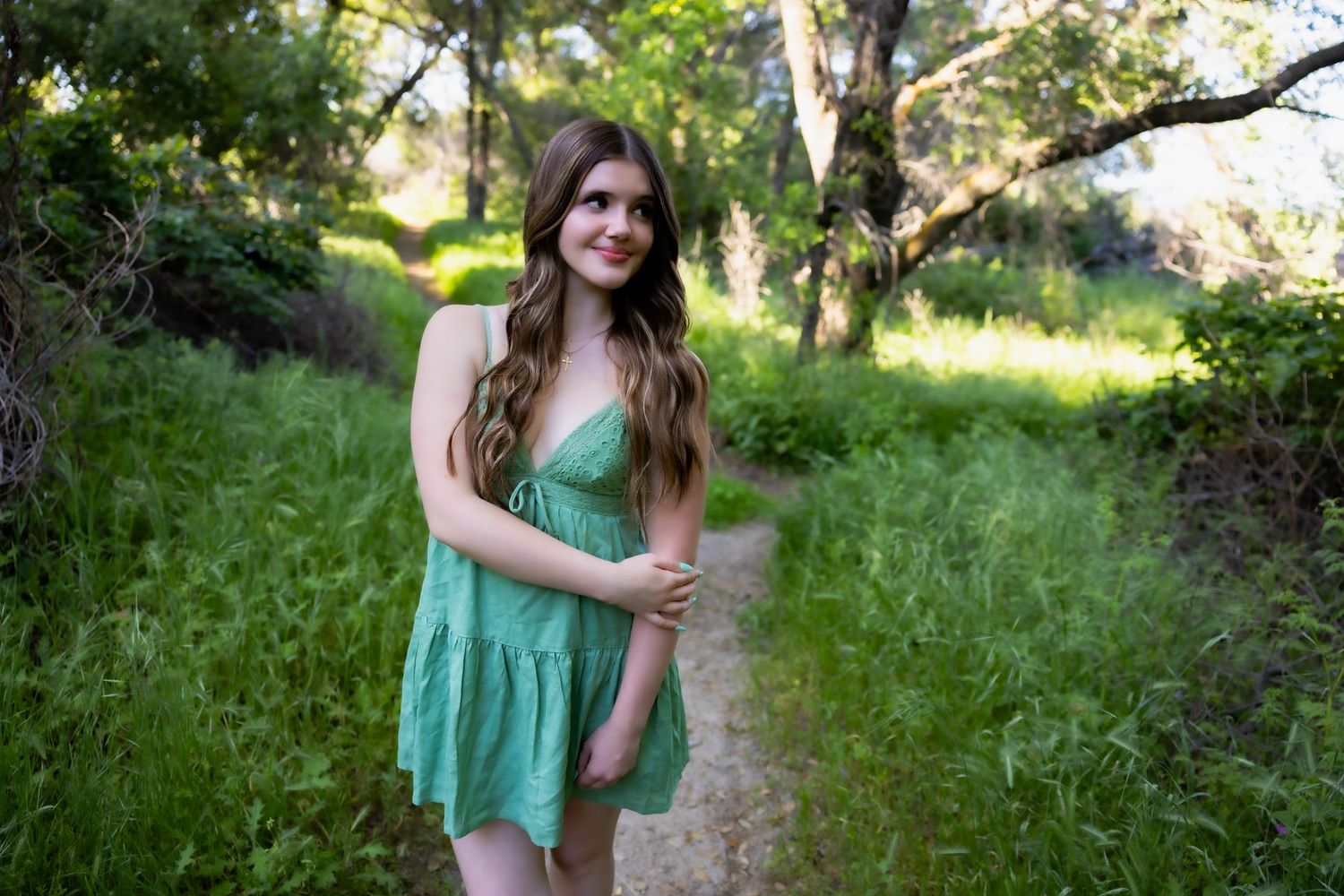 A person in a green summer dress poses along a sunlit wooded path surrounded by lush greenery.