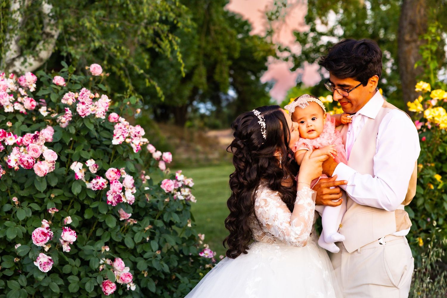 A couple shares an intimate moment outdoors in a garden with blooming pink flowers and lush greenery.