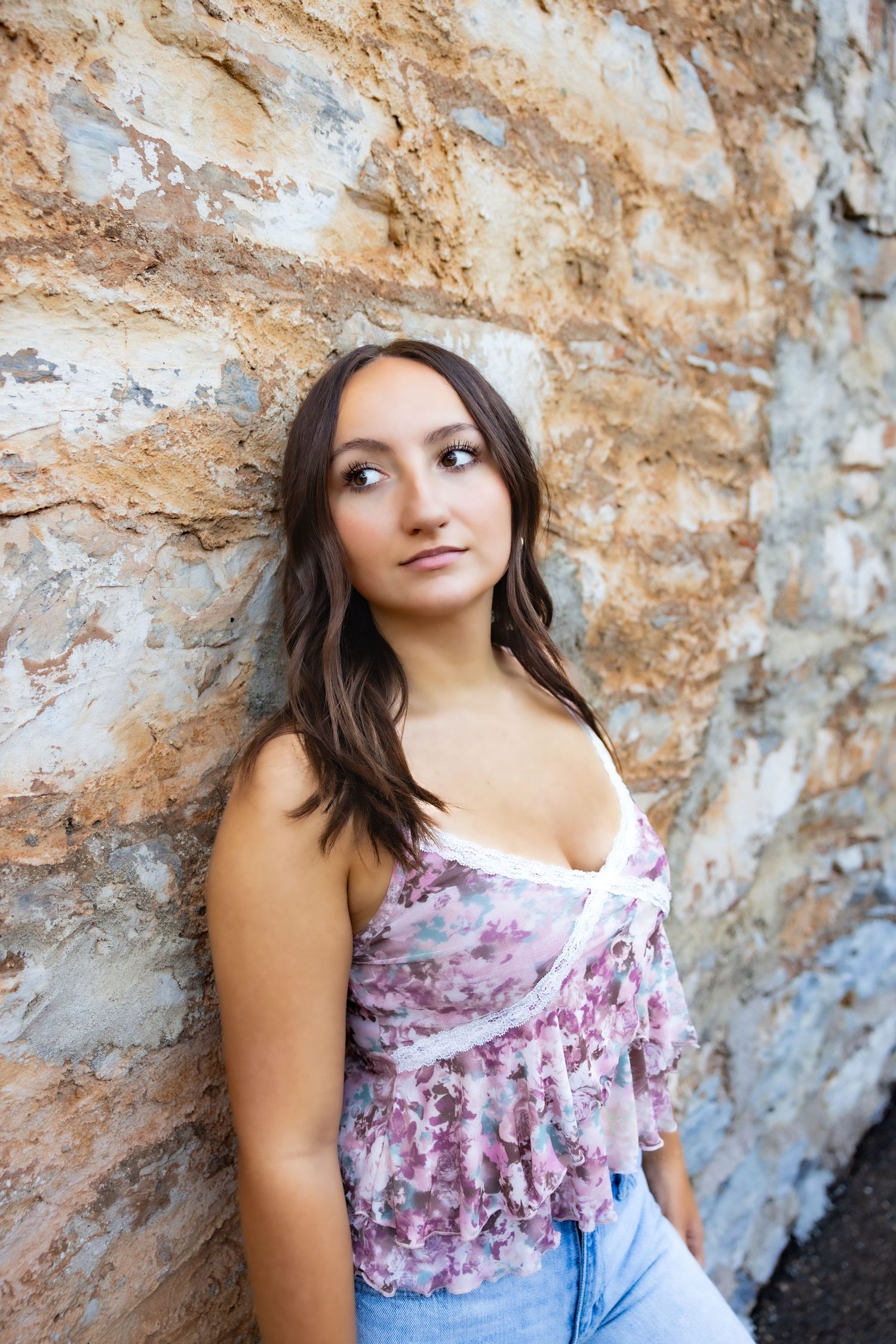 A Senior portrait against a textured stone wall wearing a floral top and jeans in Murphys, Gold Country, Amador County, Calaveras County.