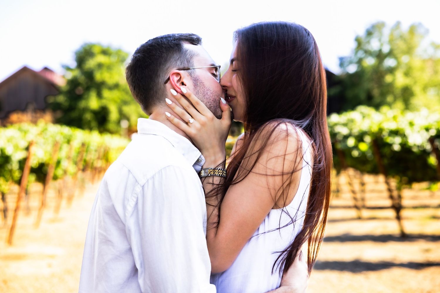 A couple shares a romantic kiss in a sunny vineyard with rows of grapevines stretching into the distance.
