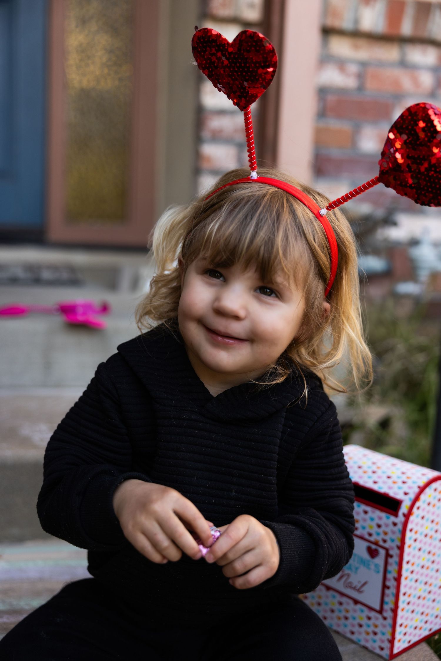 Child wearing a black sweater and sparkly red heart headband poses with a wrapped gift box during holiday season.