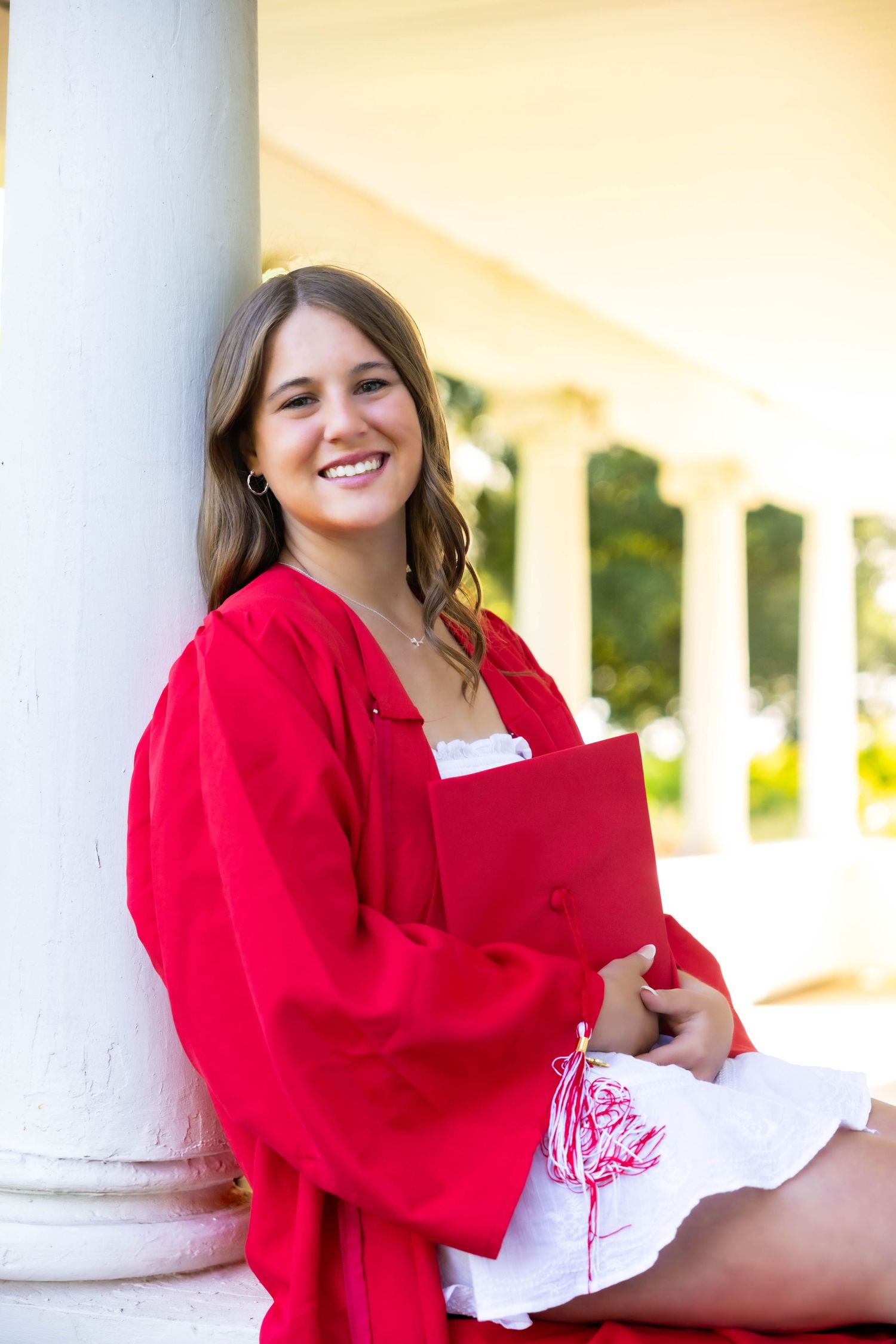 A graduate in a vibrant red gown sits by a white column in an outdoor setting holding academic materials.