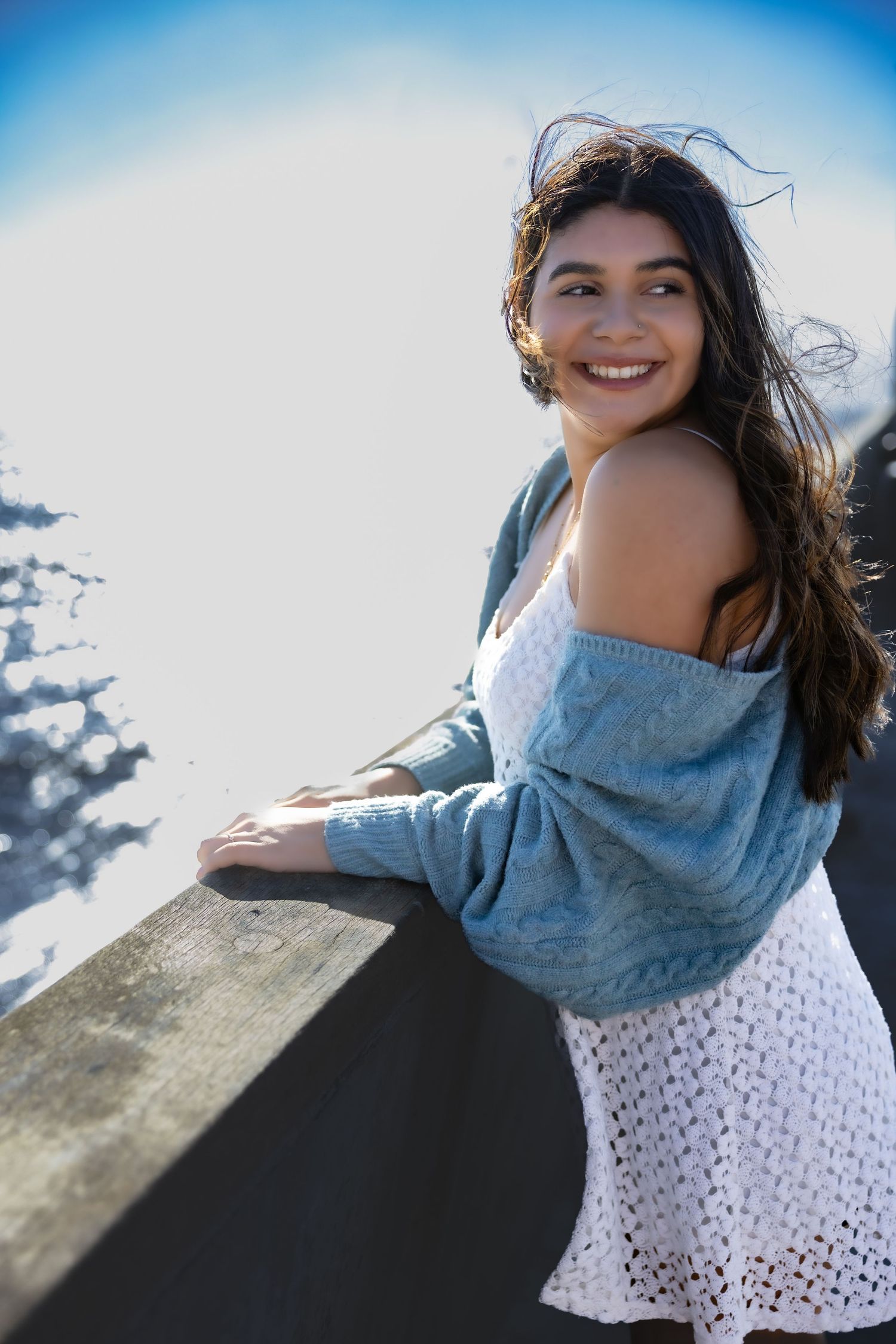 Person in an off-shoulder blue sweater and white eyelet dress standing at a scenic mountain viewpoint on a sunny day.