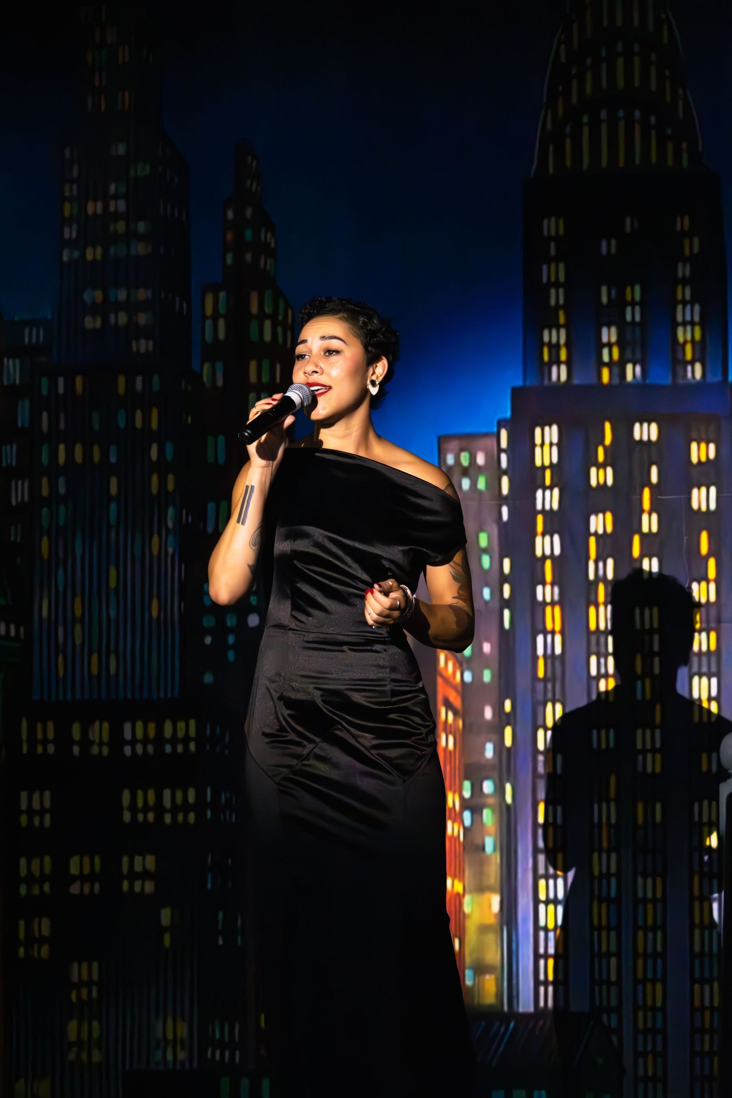 Solo performer in black outfit singing on stage with illuminated city skyline backdrop.