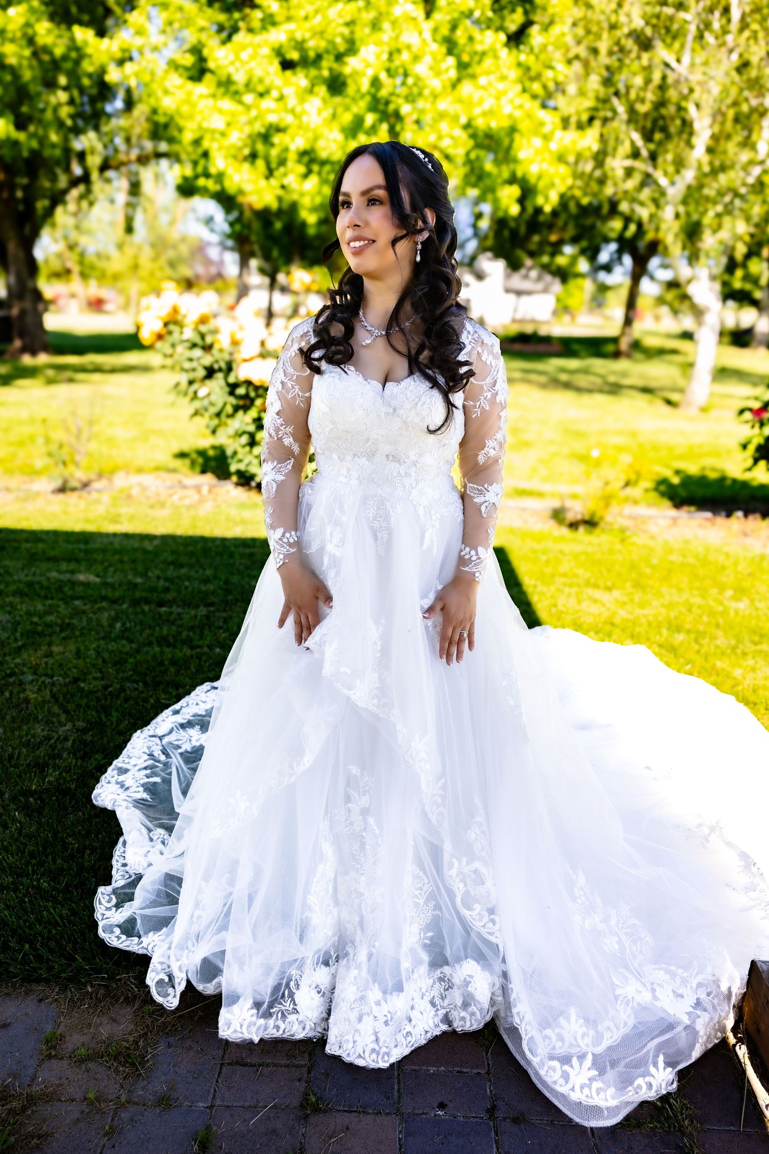 A bride in a flowing white lace gown twirls and poses outdoors in a sunny garden setting with vibrant greenery.