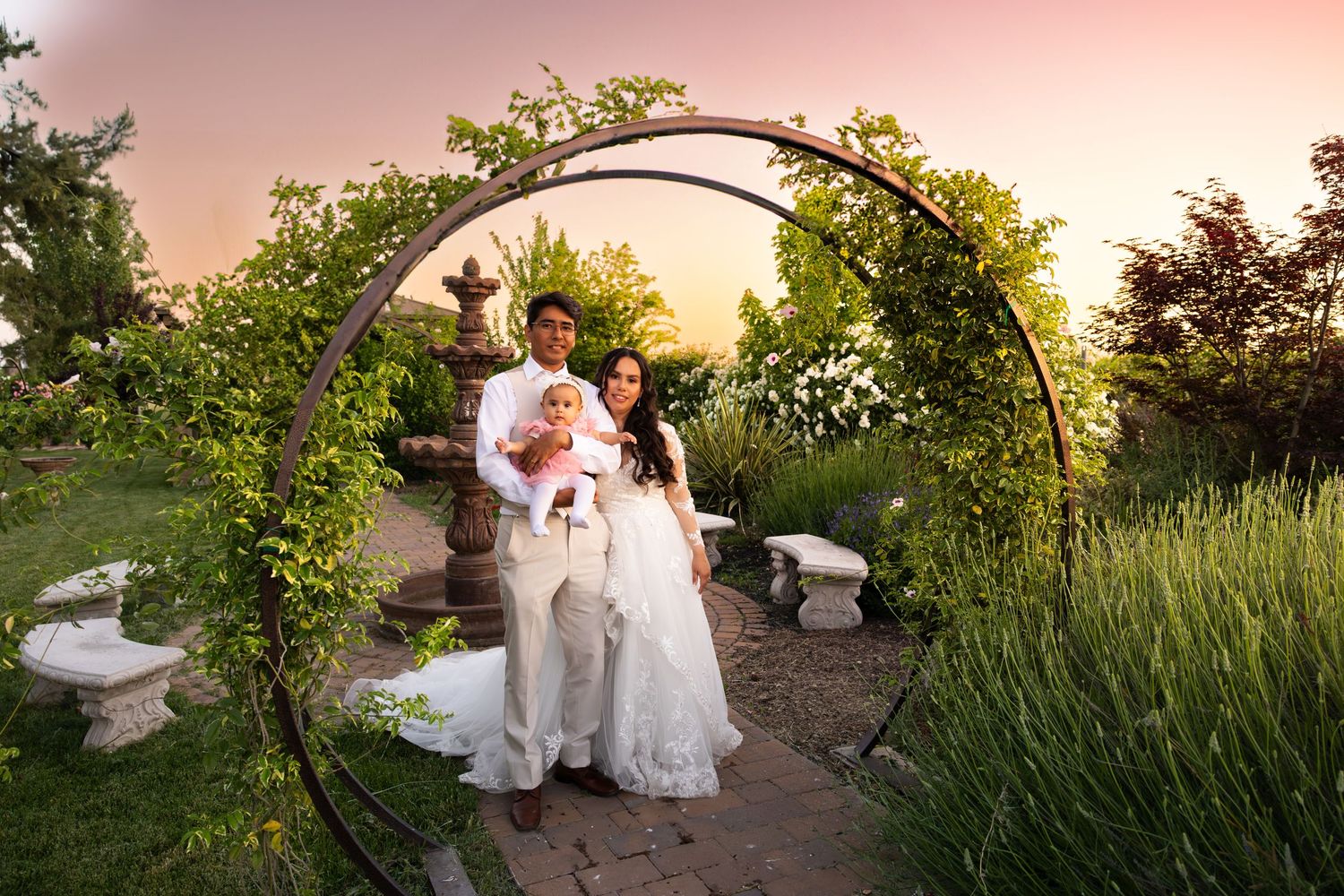 A couple walks together under a floral arch in a garden setting during sunset.