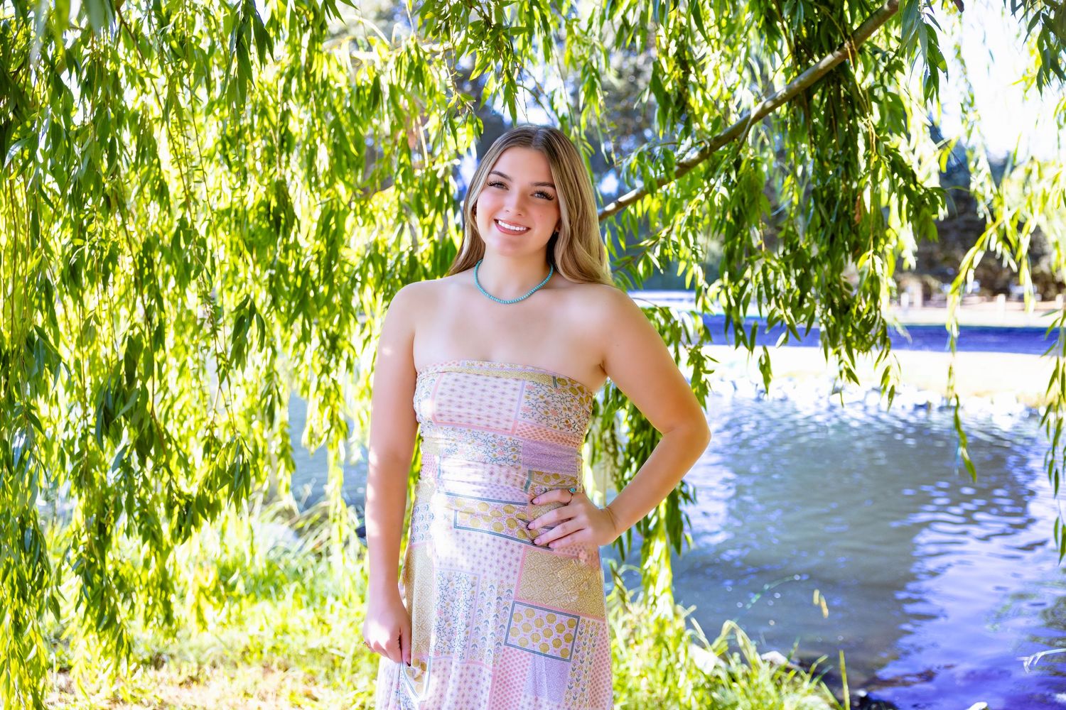 A person in a sparkly pink strapless dress poses under a weeping willow tree next to a lake.