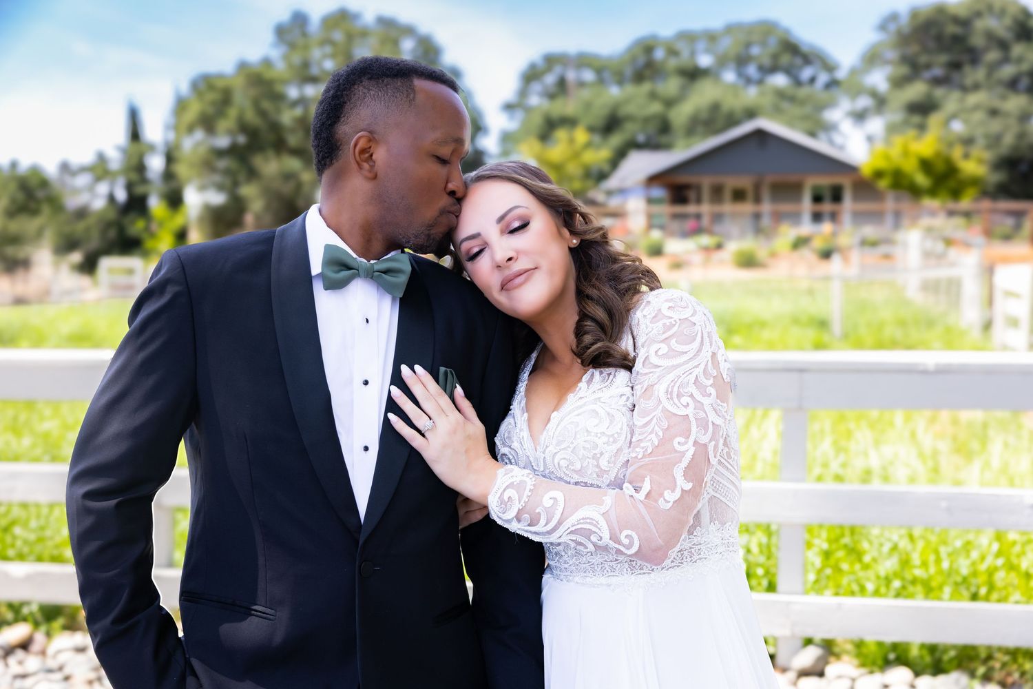 Romantic wedding portrait of a couple in formal attire embracing by a white fence on a sunny country estate.