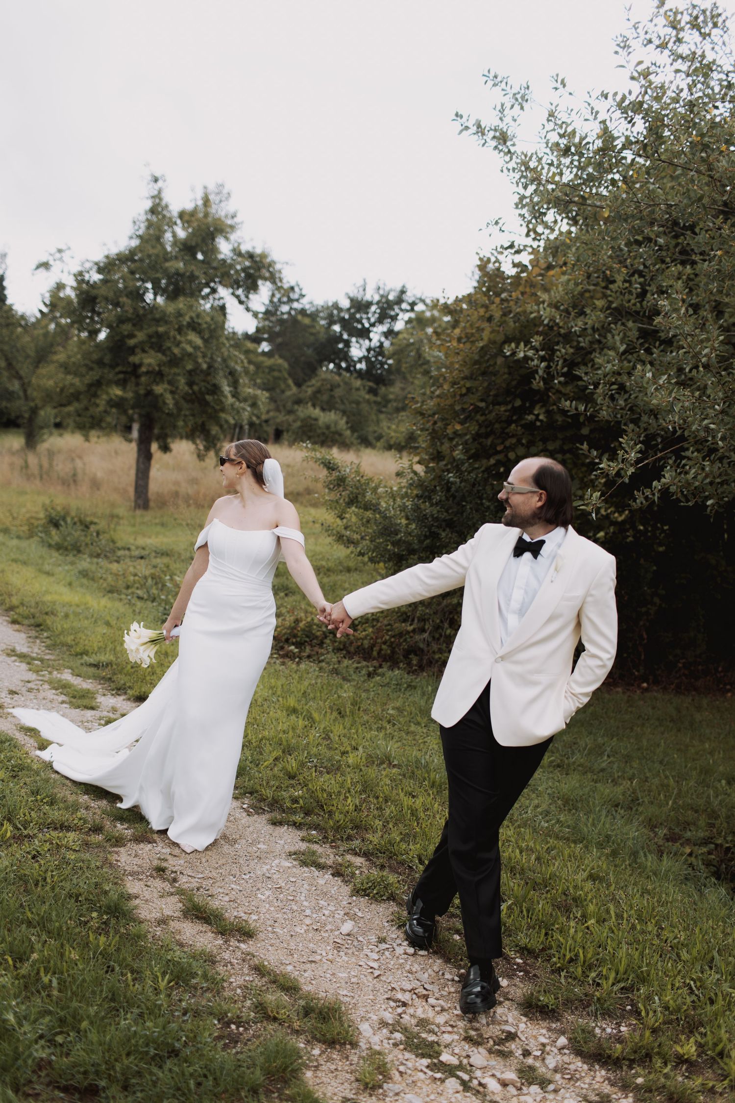 Playful wedding couple dancing and twirling on a rustic path surrounded by apple trees.